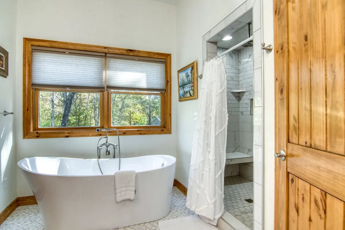 A tranquil bathroom with a freestanding soaking tub positioned in front of a large window, a separate shower area, and natural light pouring in. The clean design and spa-like details evoke a sense of relaxation and luxury.
