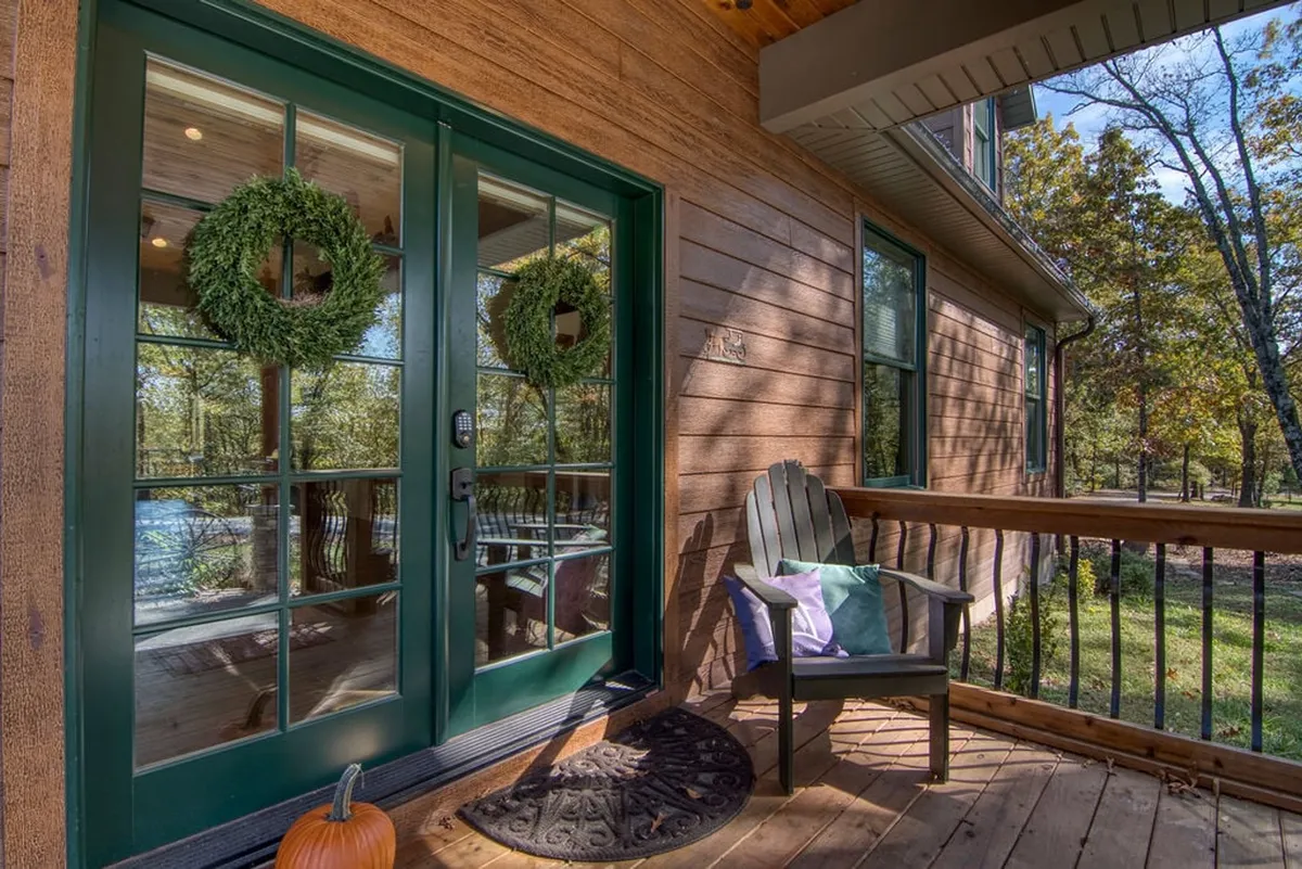 The view shows the welcoming front porch of the home, featuring inviting double green doors with seasonal wreaths, a wooden chair, and a pumpkin on the mat. The porch is shaded and overlooks the immediate outdoors, suggesting a relaxing spot to enjoy the fresh air with your pet after exploring.