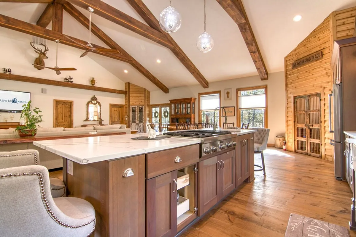 A contemporary kitchen featuring a large island with bar seating, exposed wooden beams, modern appliances, and a comfortable seating nook. The area is awash with natural light from nearby windows, making it ideal for cooking, entertaining, and socializing.