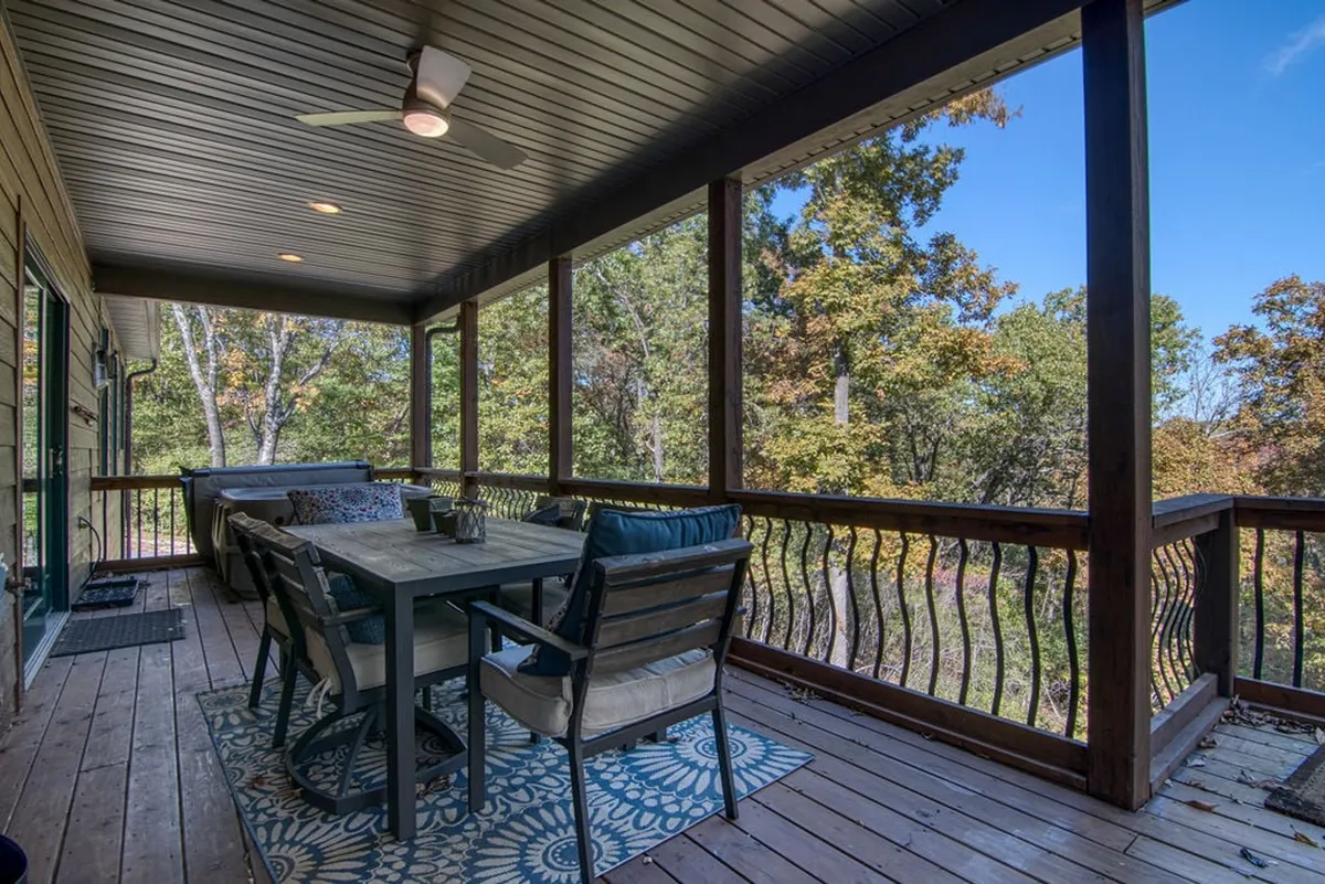 The image captures an outdoor dining table and chairs placed on a deck, shaded by a ceiling fan and surrounded by lush trees. The tranquil scene evokes the experience of alfresco dining and social gatherings beneath the open sky.
