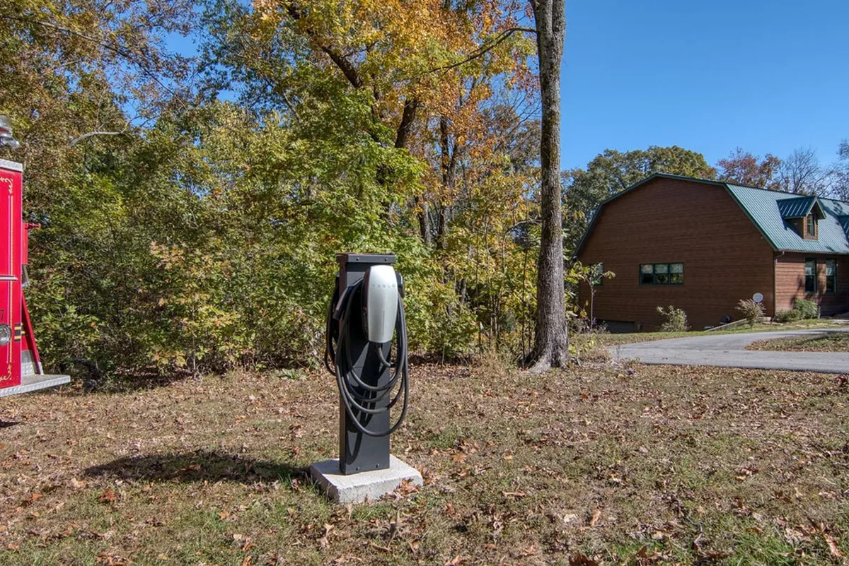The image displays an exterior scene highlighting a modern brick house surrounded by lush autumn foliage. Prominently featured is an electric vehicle (EV) charger, indicating the property's commitment to sustainability and convenience for eco-conscious travelers.
