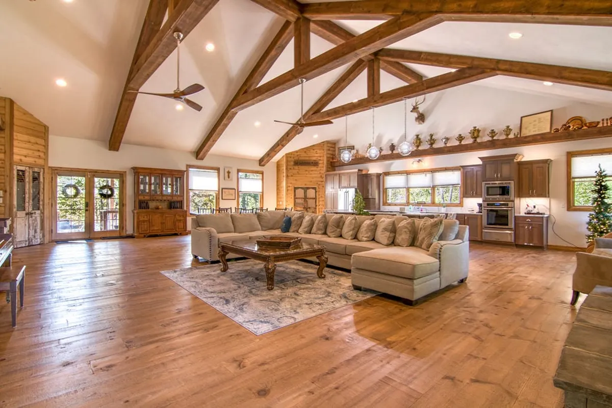The image showcases a spacious, open-concept living area with a large sectional sofa centered around a wooden coffee table, exposed wooden beams overhead, a modern kitchen in the background, and expansive windows revealing serene woodland views.