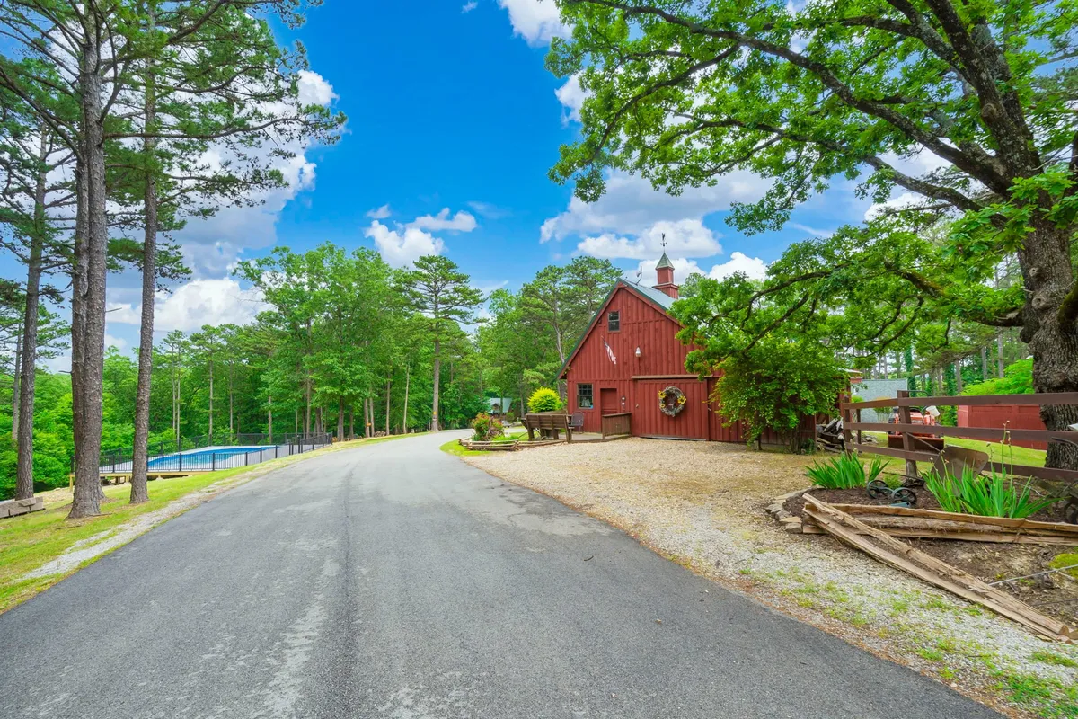 An exterior view includes a charming red barn, a gravel driveway bordered by lush greenery, and a glimpse of a swimming pool in the background. The spacious outdoor environment invites leisurely walks and outdoor play, surrounded by serene nature.