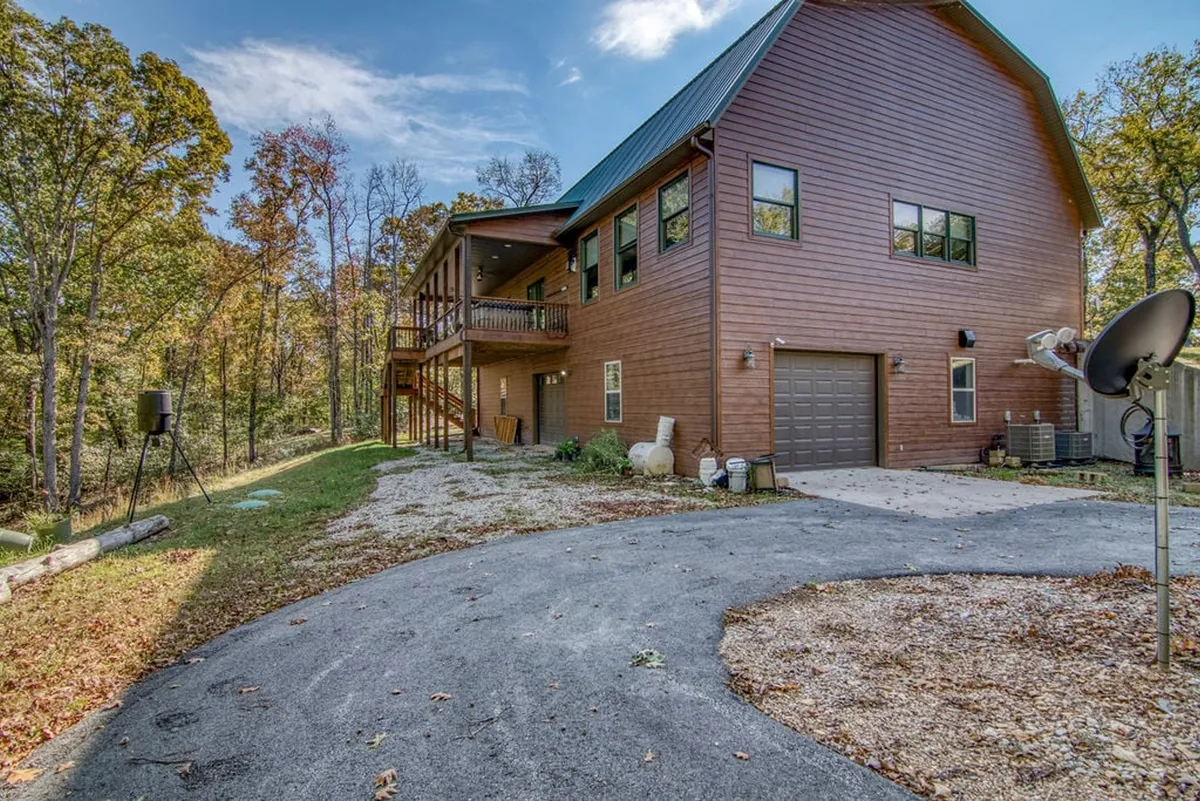 A tranquil view of a wooden house facade nestled among tall trees, with large windows and a driveway leading up to the home. The forested surroundings evoke a sense of privacy and immersion in nature.
