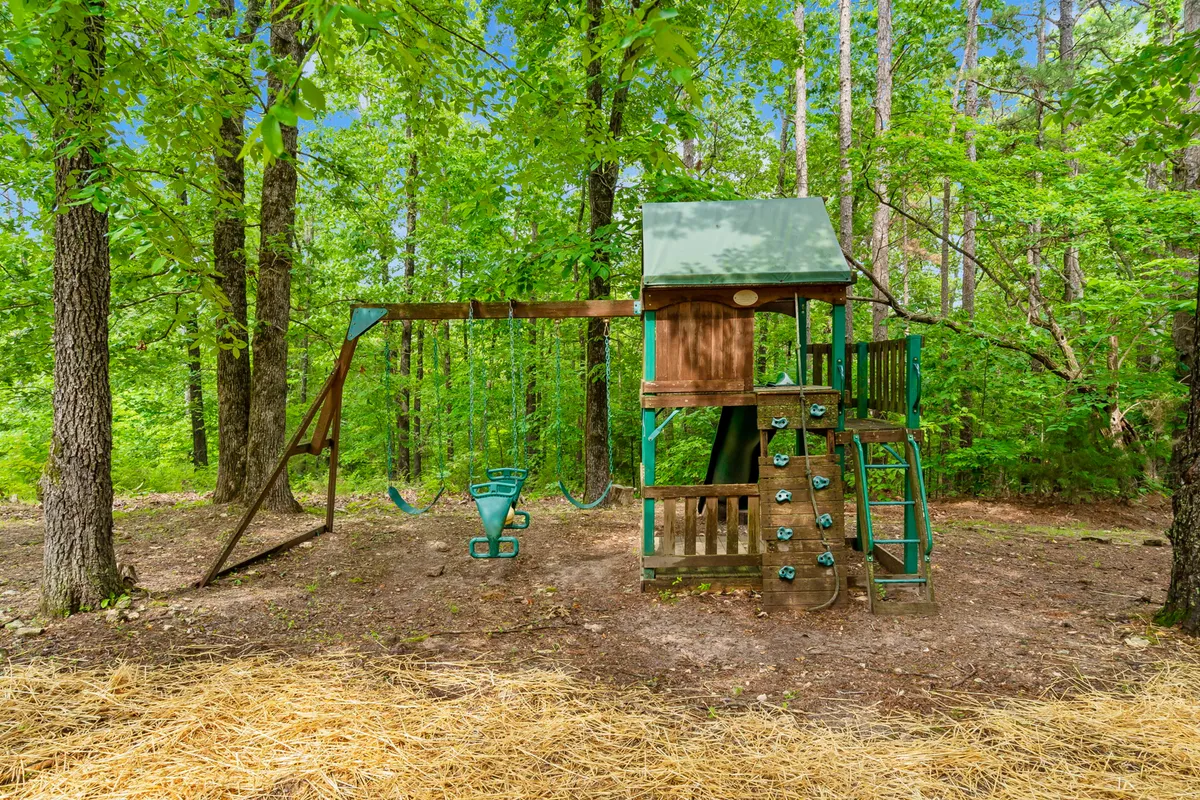 This cheerful image features a playground structure with a swing set and a climbing wall, all nestled beneath tall, leafy trees. The area is vibrant and playful, inviting families and children to enjoy the outdoors together.