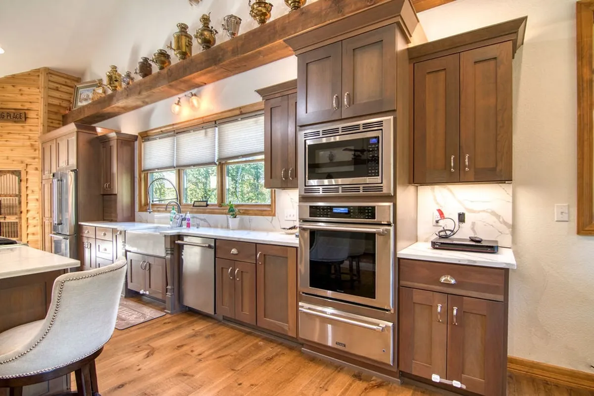 A bright, inviting kitchen featuring a quartz island as the centerpiece, surrounded by wooden cabinetry and stainless steel appliances. Large windows bathe the space in natural light, creating a warm, welcoming atmosphere. Decorative items on shelves add a touch of personality.