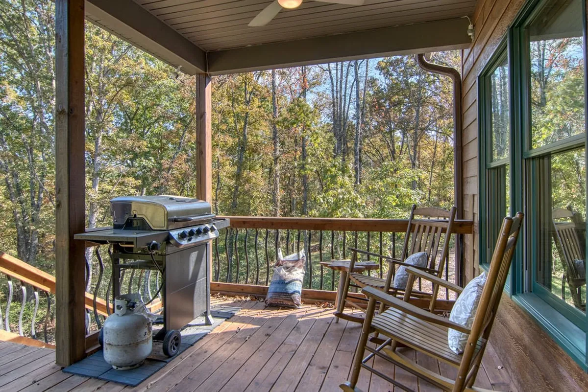 A charming wooden deck featuring a BBQ grill, rocking chairs, and lush greenery. The area invites relaxation, grilling, and informal outdoor gatherings, with sunlight filtering through the trees.