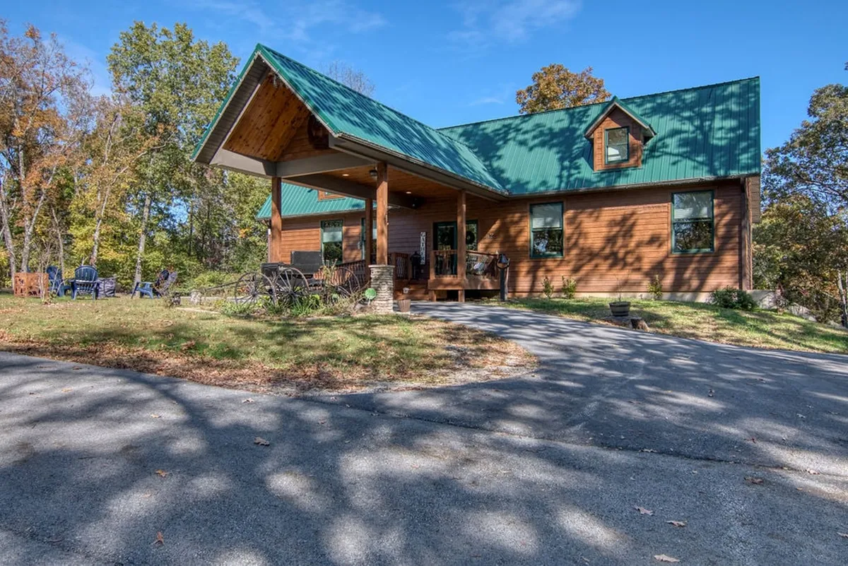 A charming wooden house with a green metal roof sits nestled among lush trees. The inviting front porch features Adirondack chairs, perfect for relaxing and soaking in the peaceful natural surroundings.