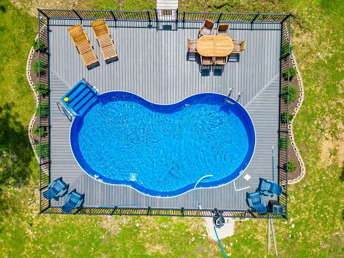 This image showcases a sparkling swimming pool with a deck area, outdoor dining table, and lounge chairs surrounded by greenery. The scene is set up for socializing, swimming, sunbathing, and enjoying outdoor meals together.