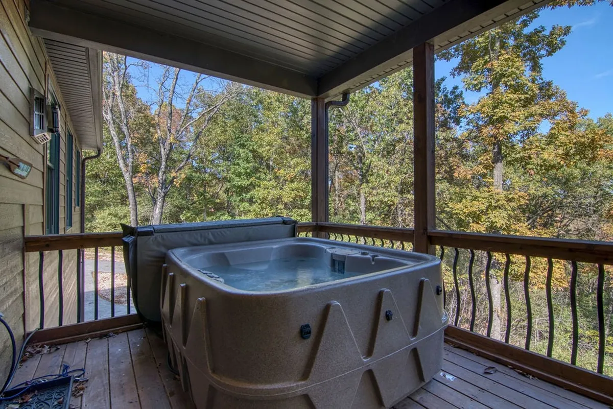 This image showcases a tranquil wooden deck nestled among tall trees, featuring a bubbling hot tub as its focal point. The hot tub is situated for direct views into the peaceful, green woods, creating an inviting atmosphere for relaxation and nature immersion.