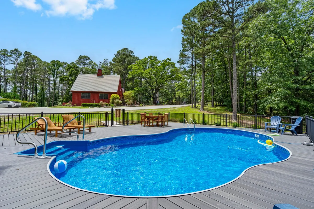 A relaxed scene featuring a community saltwater swimming pool, set on a wooden deck beside a red house and surrounded by green trees. Lounge chairs and the inviting pool highlight opportunities for relaxation and recreation.