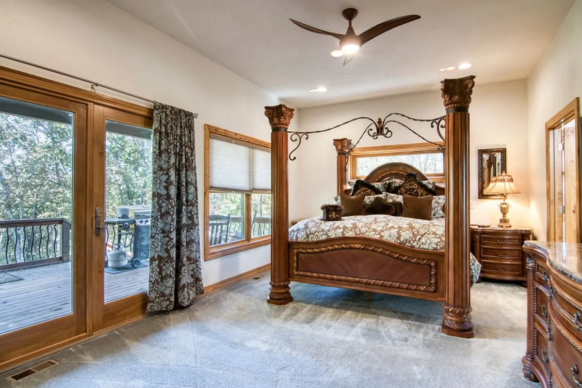 A serene bedroom featuring a four-poster bed, large windows draped with decorative curtains, and a nightstand with a lamp. The soft lighting and tranquil décor create an inviting atmosphere, while the windows offer woodland views.